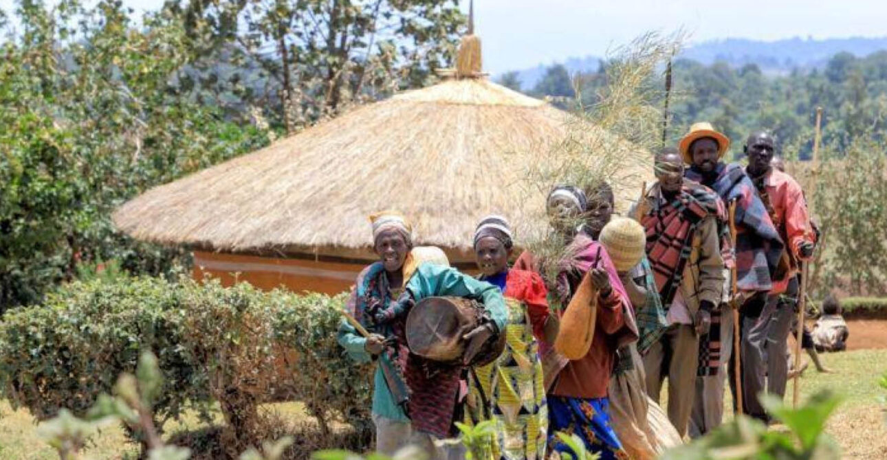 Community members in traditional clothing walking near a thatched cultural homestead