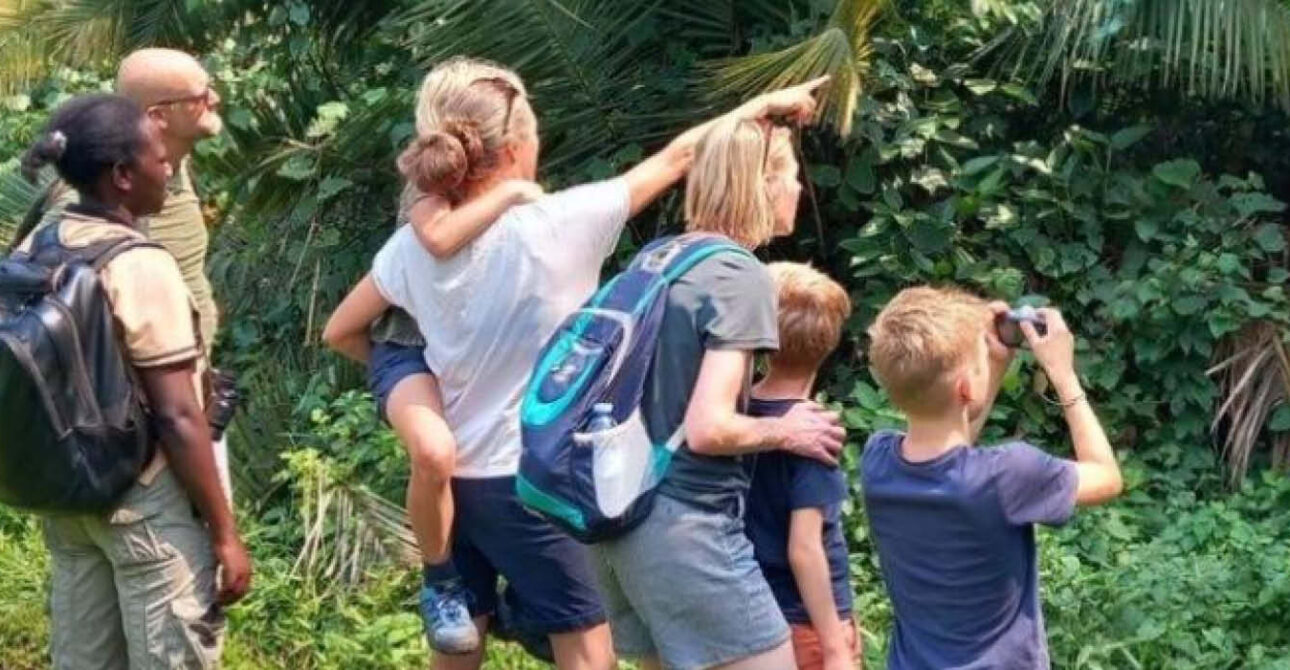 Family with children of different ages exploring a tropical forest with a guide, while one child observes nature through binoculars.