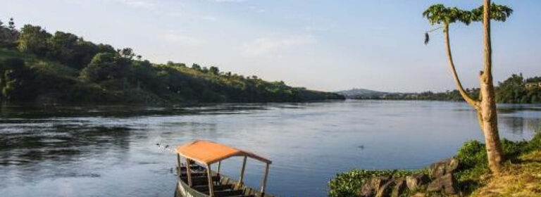 Scenic view of the River Nile in Jinja, Uganda, with a wooden boat along the riverbank, lush greenery and a clear blue sky.