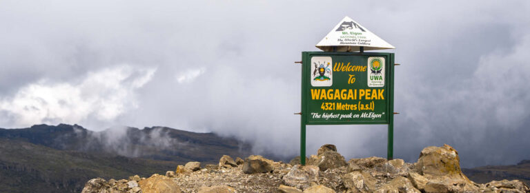 Wagagai Peak summit sign at 4,321 metres in Mount Elgon National Park, Uganda, surrounded by rocky mountain scenery and clouds.