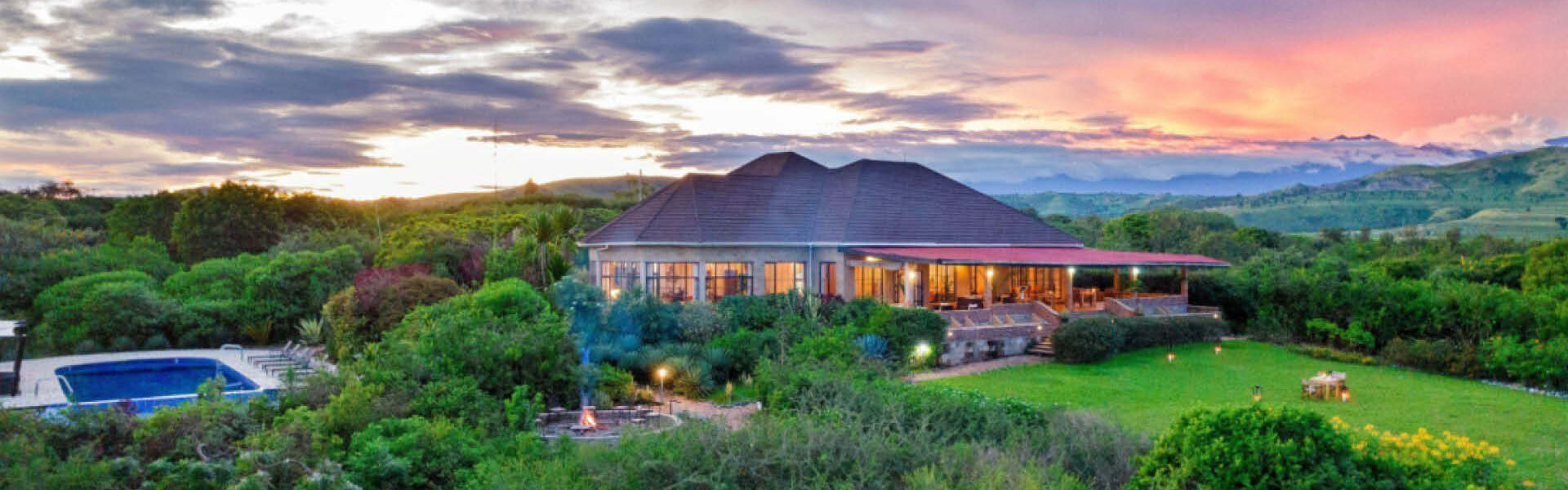 Safari lodge surrounded by lush vegetation, with a swimming pool and outdoor terraces overlooking distant hills at sunset.