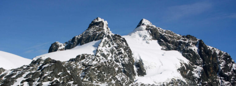 Snow-covered Margherita Peak and Alexandra Peak in the Rwenzori Mountains, Uganda, surrounded by rugged rock formations and mountain clouds.