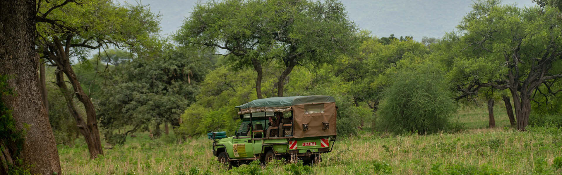 A Uganda Safari Company operating an open-sided safari vehicle on a wildlife game drive through a wooded savannah landscape in Uganda.