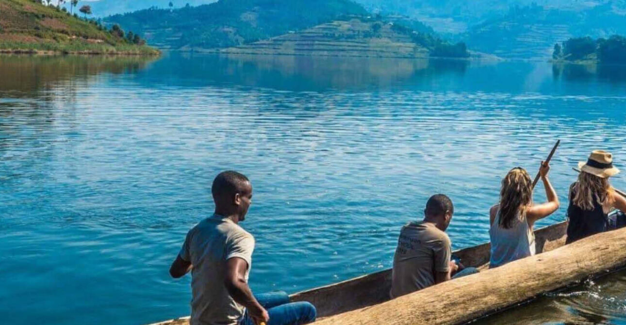 Four travellers paddling a traditional wooden canoe across a scenic lake surrounded by green hills